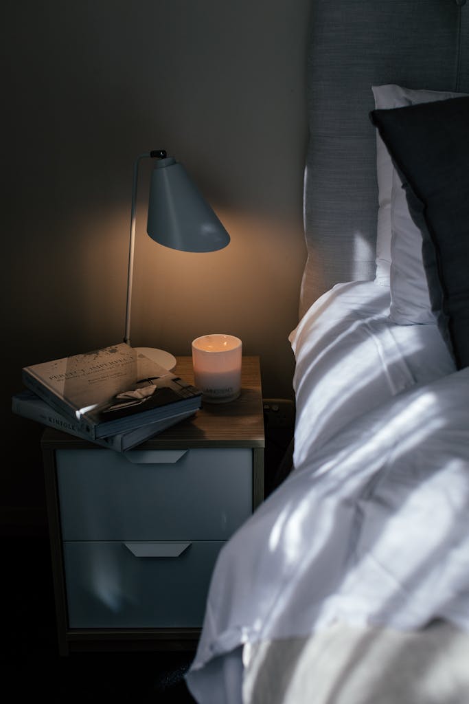 Cozy bed with crumpled sheet and pillows near nightstand with textbooks and lamp in bedroom